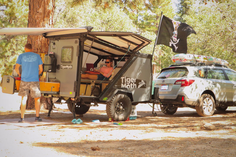 a Subaru pulling a small camper, a man cooking behind it while a woman works on her laptop within
