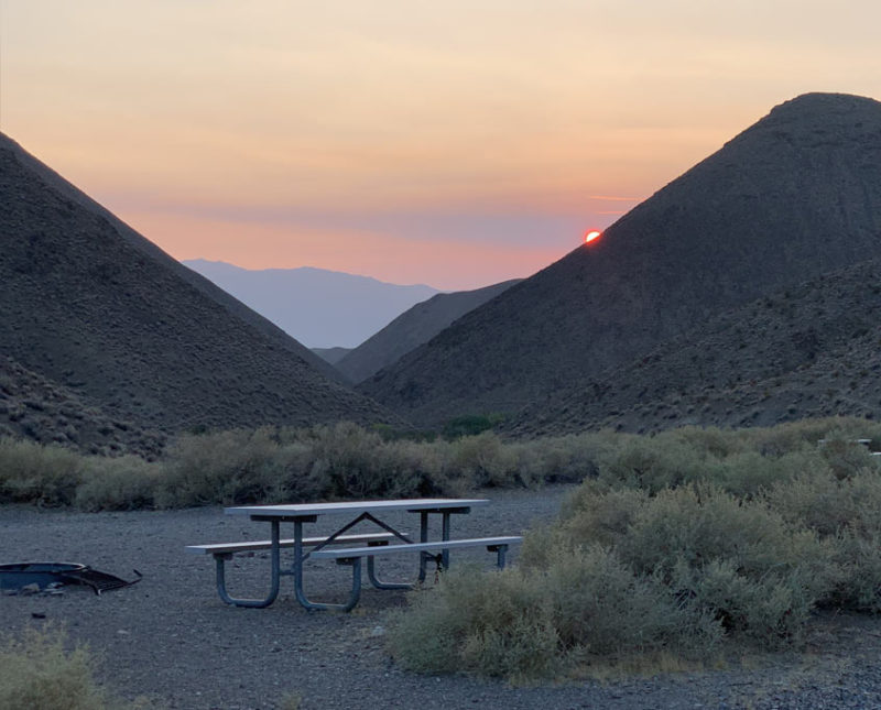 a picnic table and fire ring at a campsite, beneath tall mountains and the setting sun over Death Valley National Park