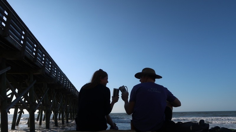 the silhouettes of two people sitting near a dock by the shore clinking coffee cups