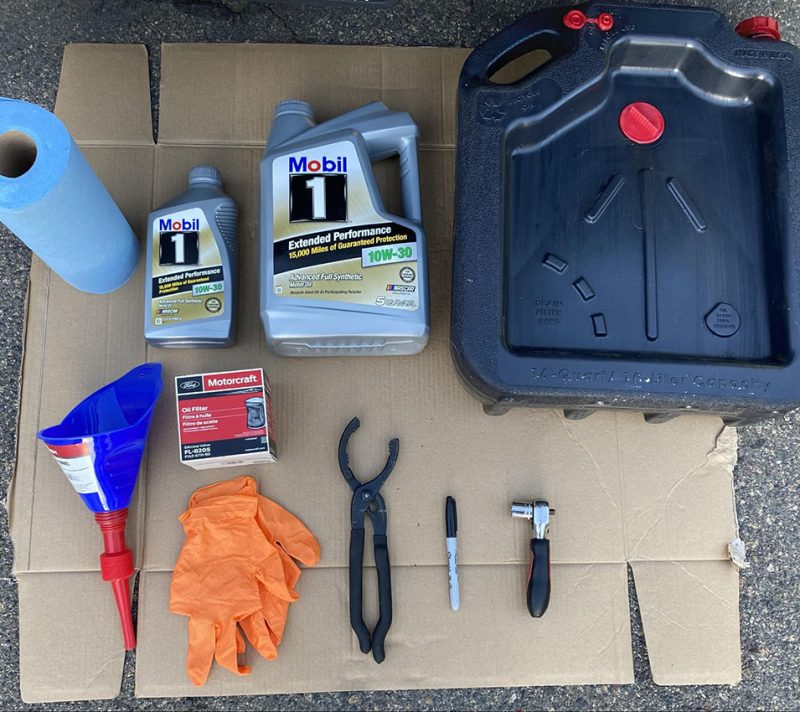 A roll of paper towels, 2 jugs of oil, an oil pan, a funnel and gloves laid out on cardboard