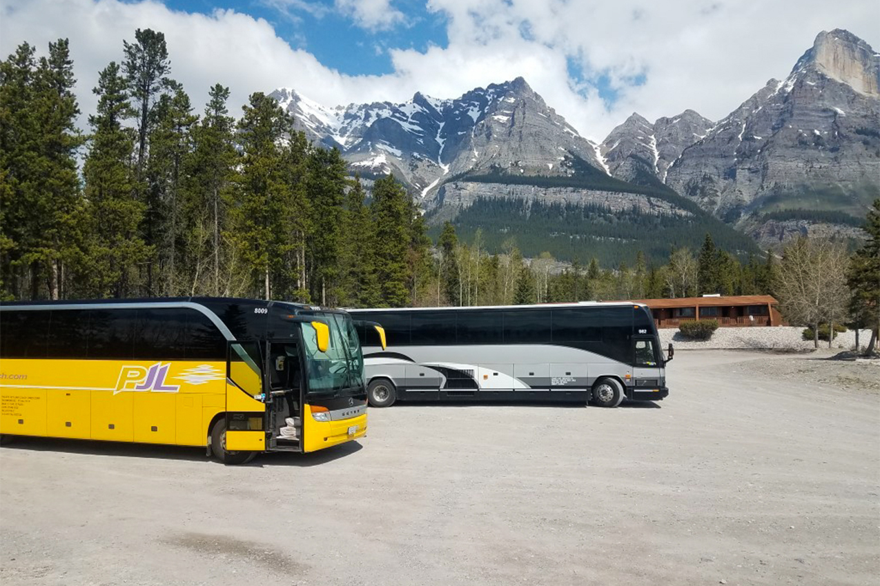Two large buses parked in front of trees and mountains.