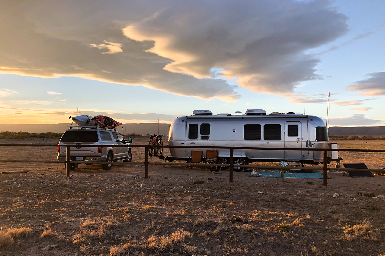 Airstream and truck with kayaks on the roof boondocked in a gravel lot at sunset.