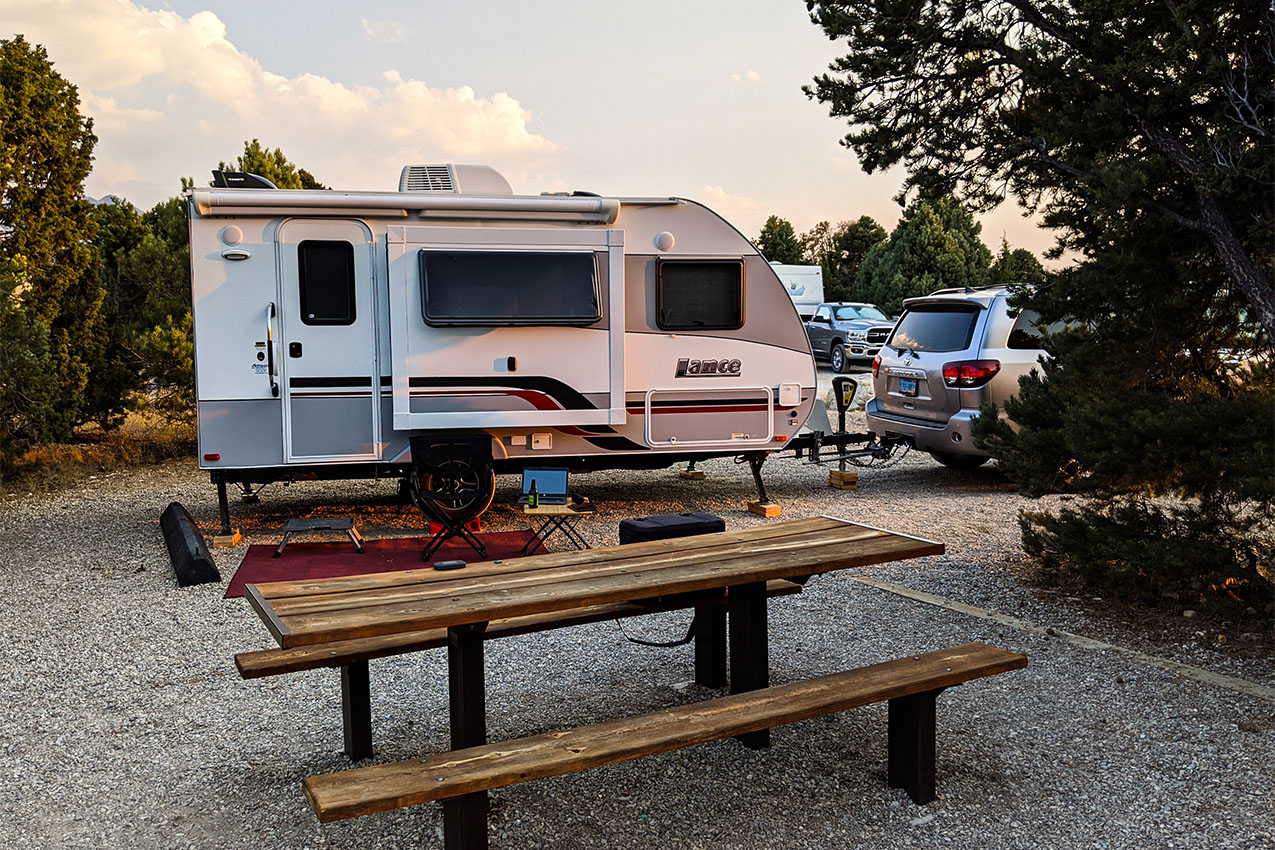 Travel trailer and car parked in a campsite next to a picnic table.