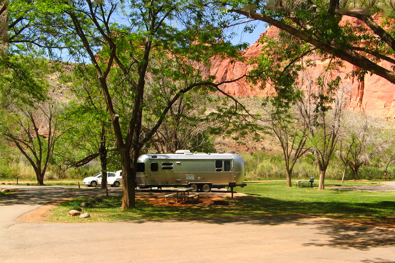 Airstream parked under trees and surrounded by red rocks.