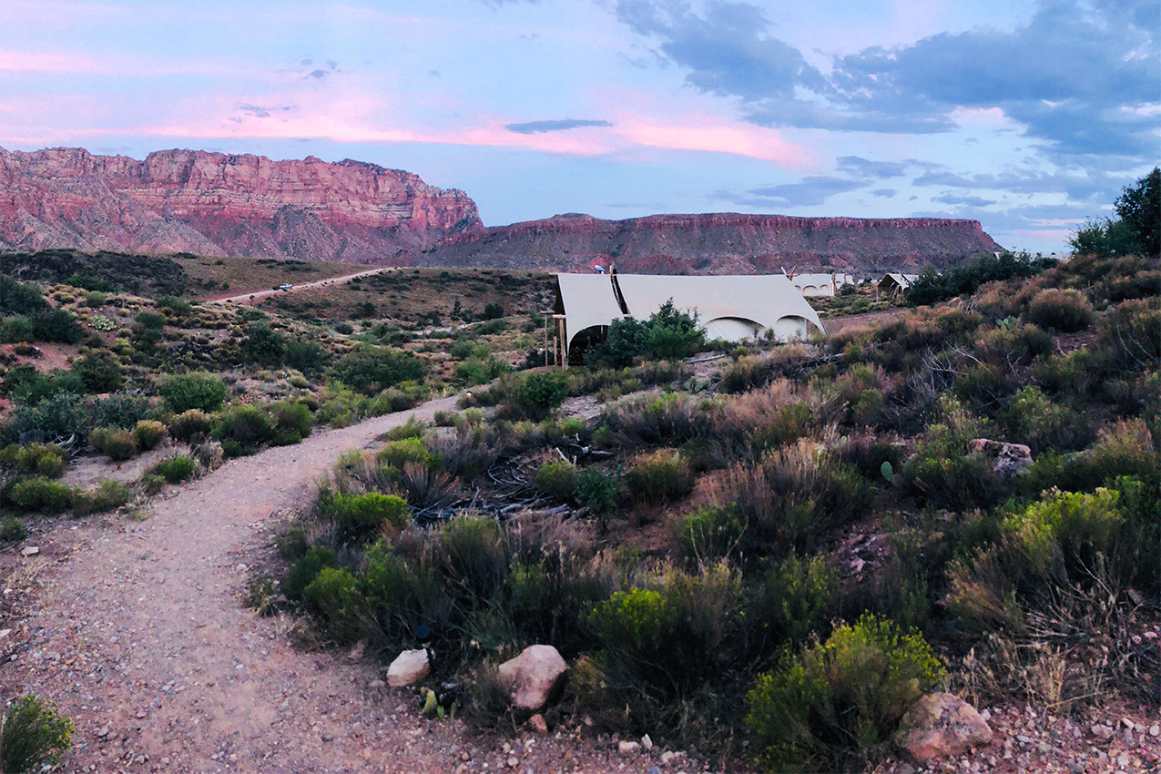 Rock path leading to a canvas tent surrounded by shrub and red mountains and pink sky.