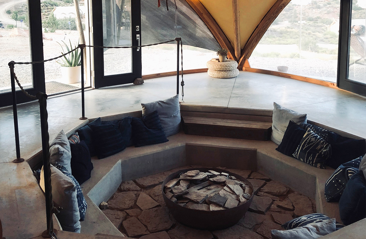 Concrete seating area filled with pillows inside a windowed canvas tent with a fire pit in the middle.