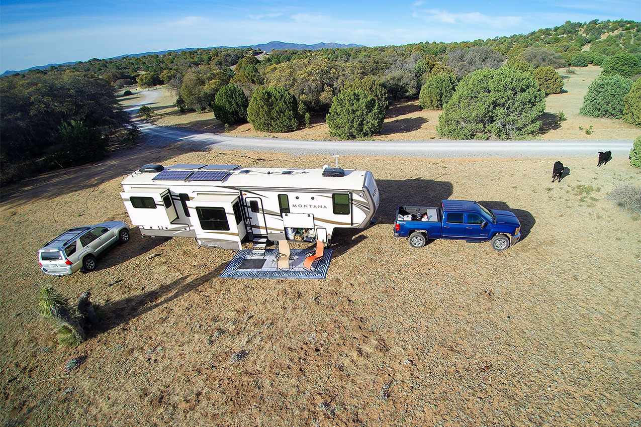 Aerial photo of a large fifth wheel and two vehicles parked in an open field surrounded by juniper bushes.