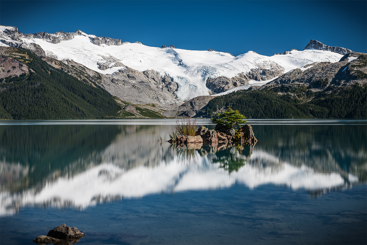 Snow covered mountains reflected in a lake with a small island in the middle.