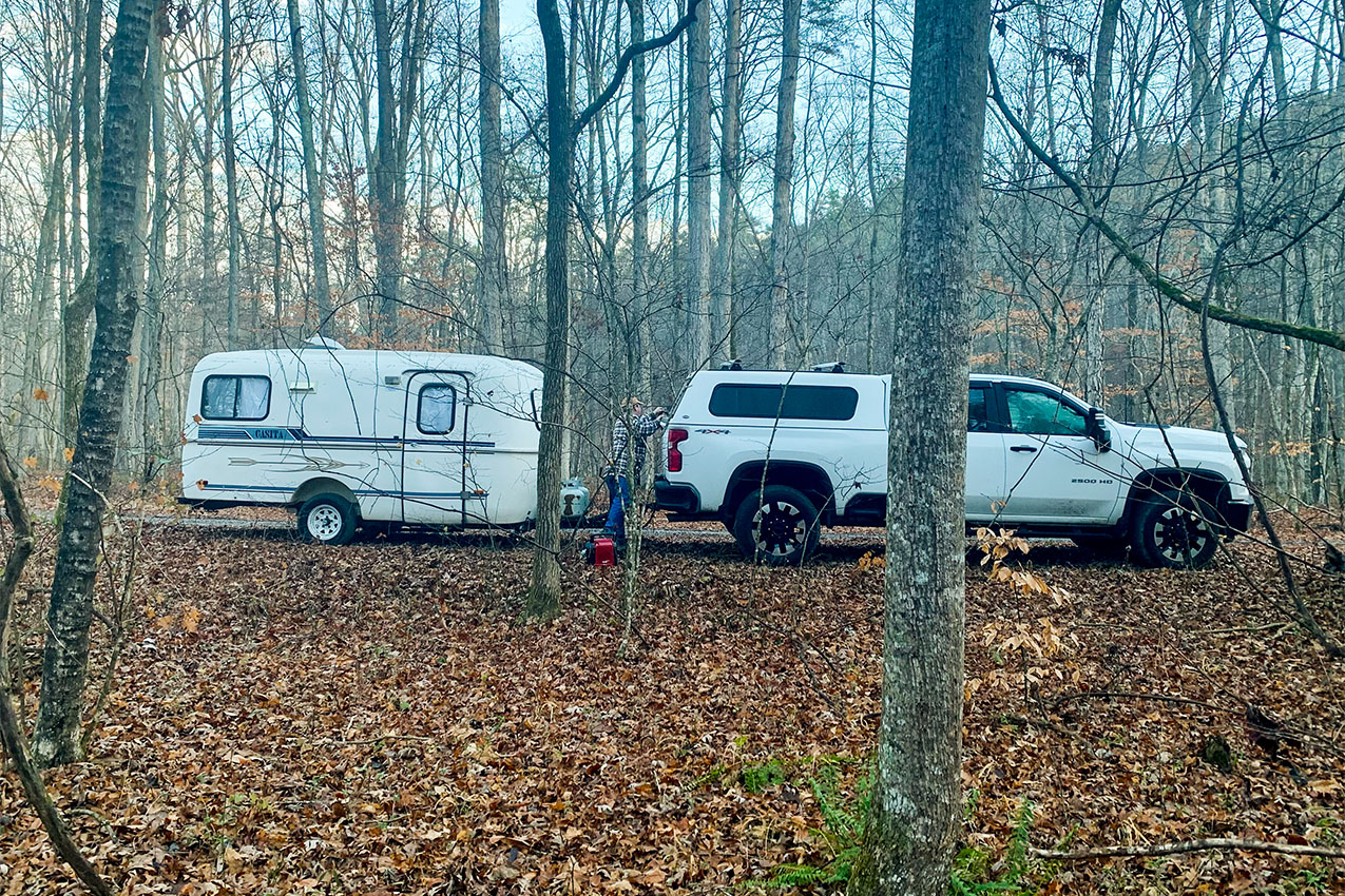Truck and small travel trailer parked in dense woods covered in fallen leaves.