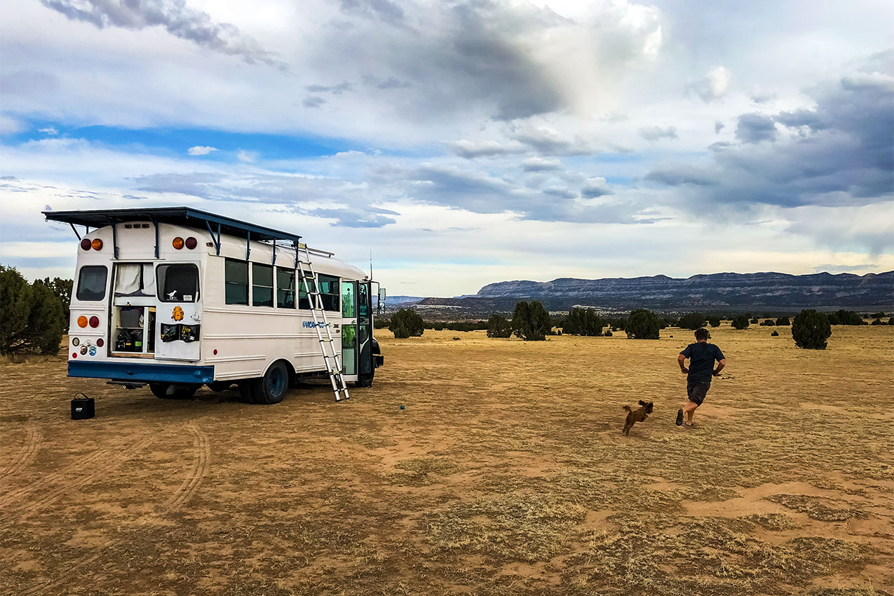A guy and his dog running next to their converted school bus in the desert.