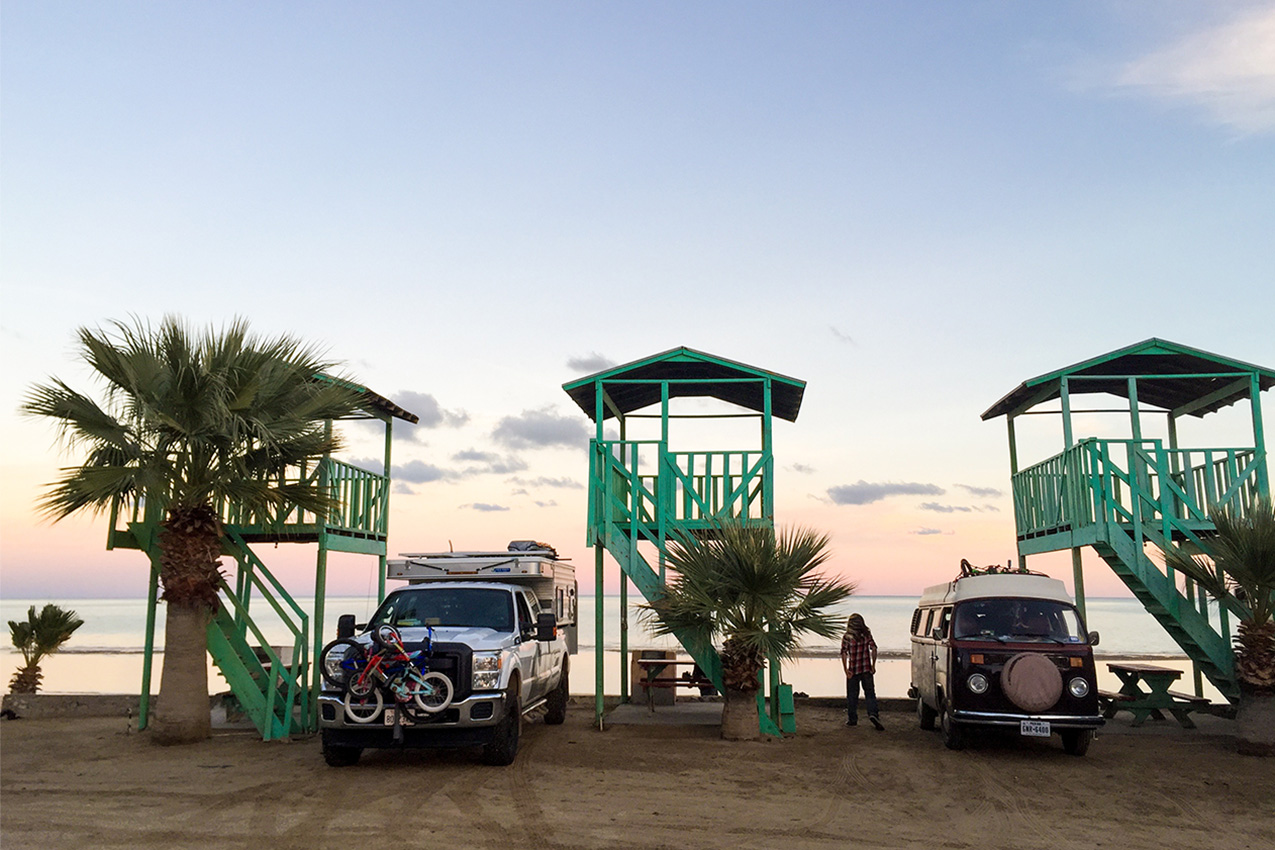 Green palapas line a beach with palm trees, truck camper, and a Westy.