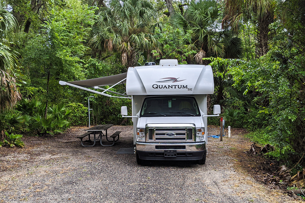 RV with awning out parked under a canopy of trees.