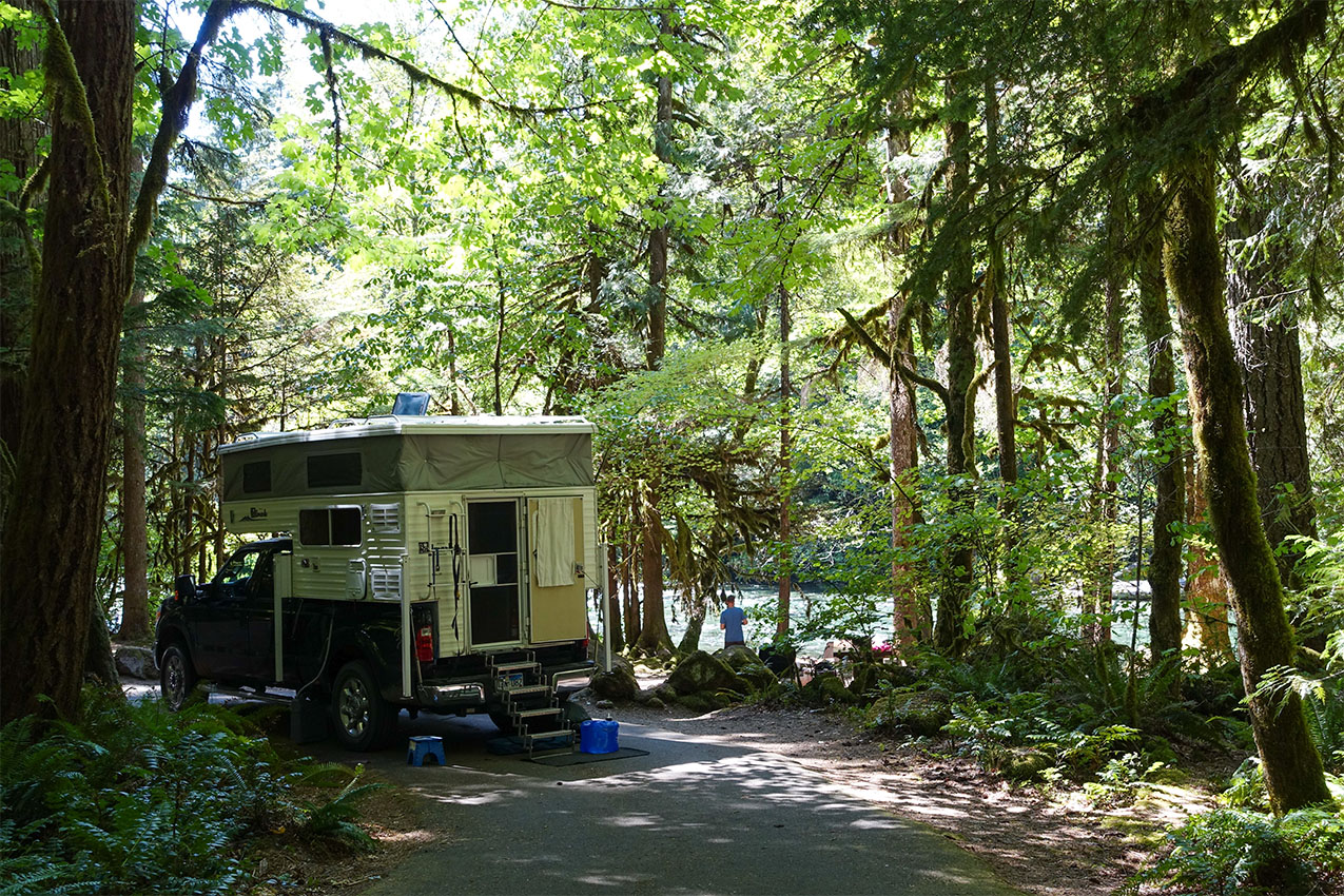 Truck camper parked under dense forest next to a river.