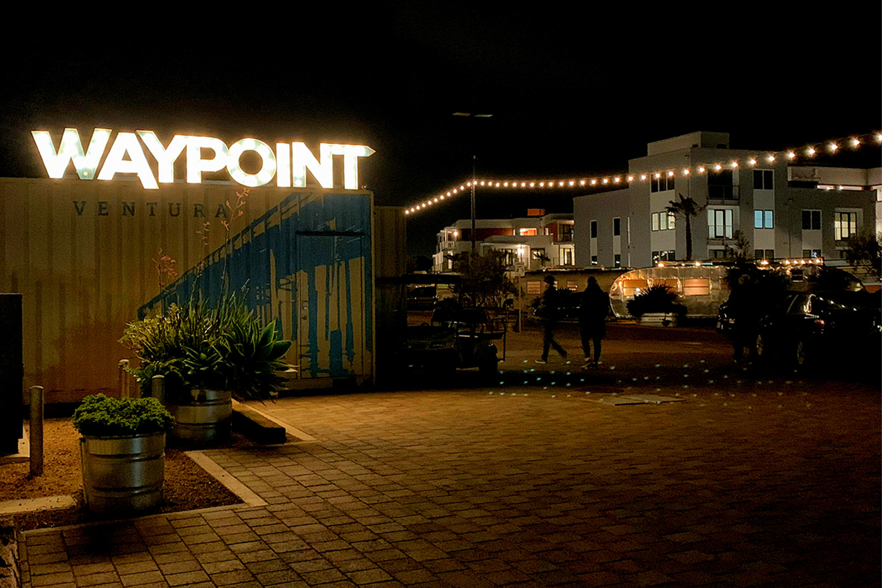 Night photo of lit "Waypoint" sign with string lights and people walking next to an Airstream.