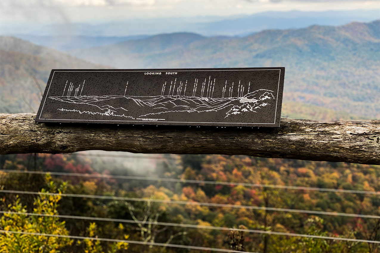 Metal sign with names of mountain peaks installed on a handrail overlooking fall colored mountains.