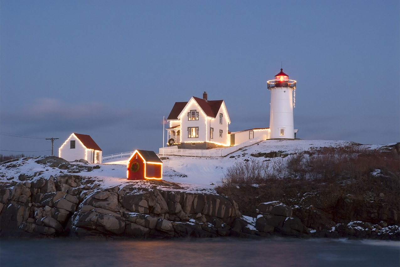 Nubble lighthouse lit up in white Christmas lights at dusk during the holidays on the coast of southern Maine.