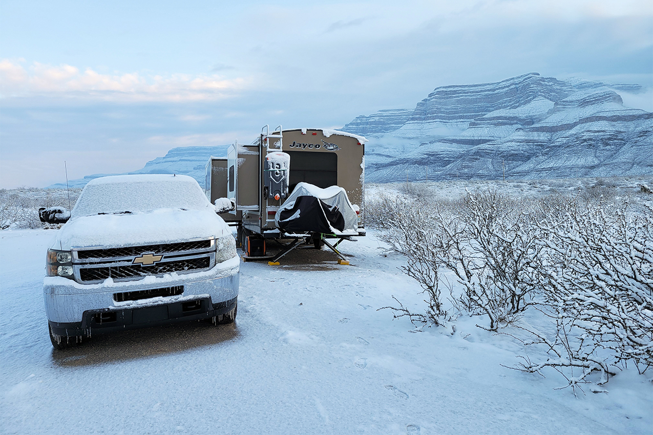 Truck and travel trailer covered in snow parked in a high desert with mountain in the back.