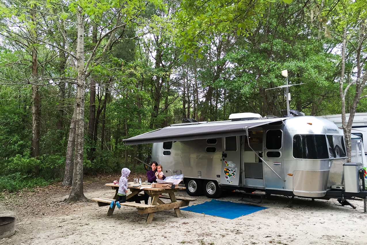 Woman with two kids sitting at a picnic table next to their Airstream in the woods.