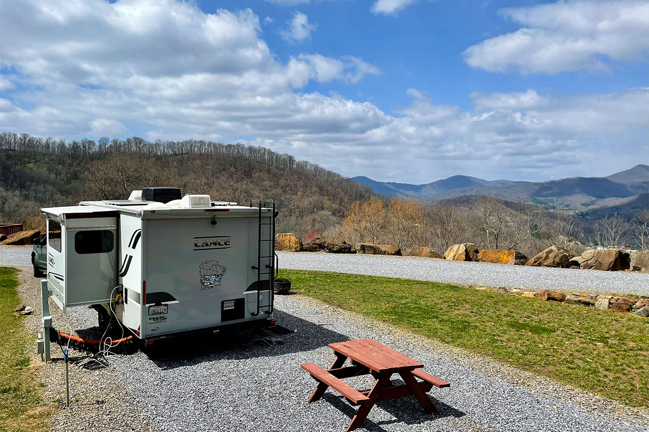 RV with slide out parked in a gravel site in front of a picnic table overlooking mountains.
