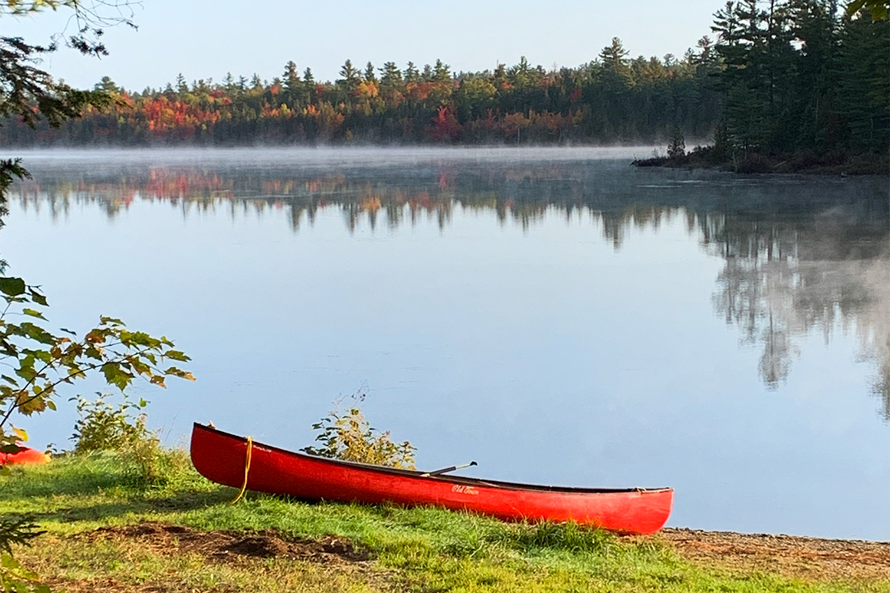 Red canoe sitting on the shore of a lake surrounded by trees.