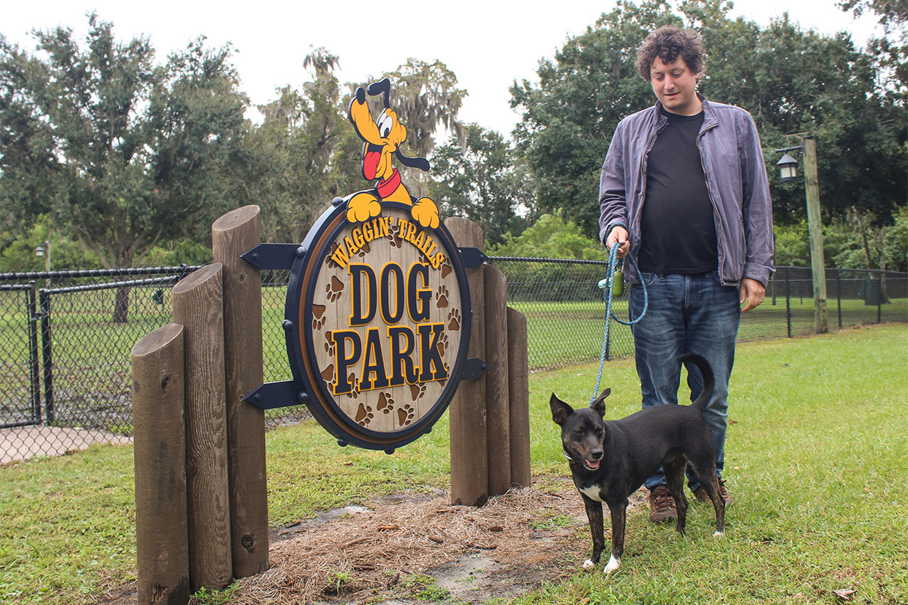 Man and dog standing in front of wooden dog park sign.