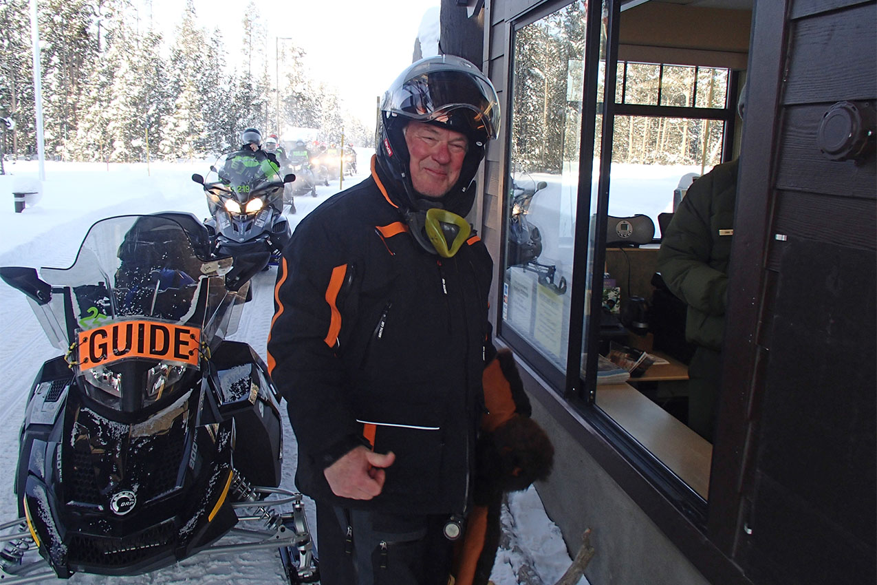 Man in winter gear standing at national park entrance booth in front of his snowmobile.