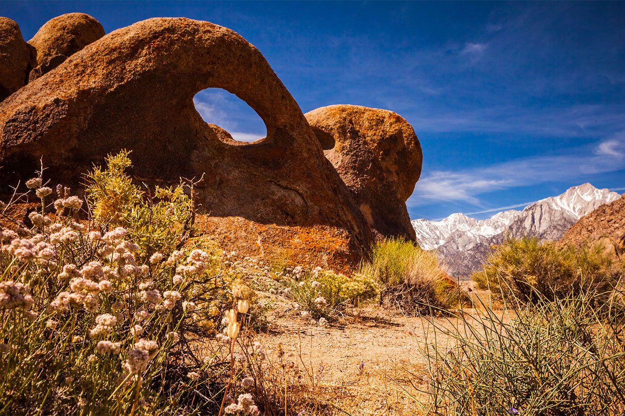 A rounded rock formation with a large hole in it under blue skies with snowcap mountains in the background.