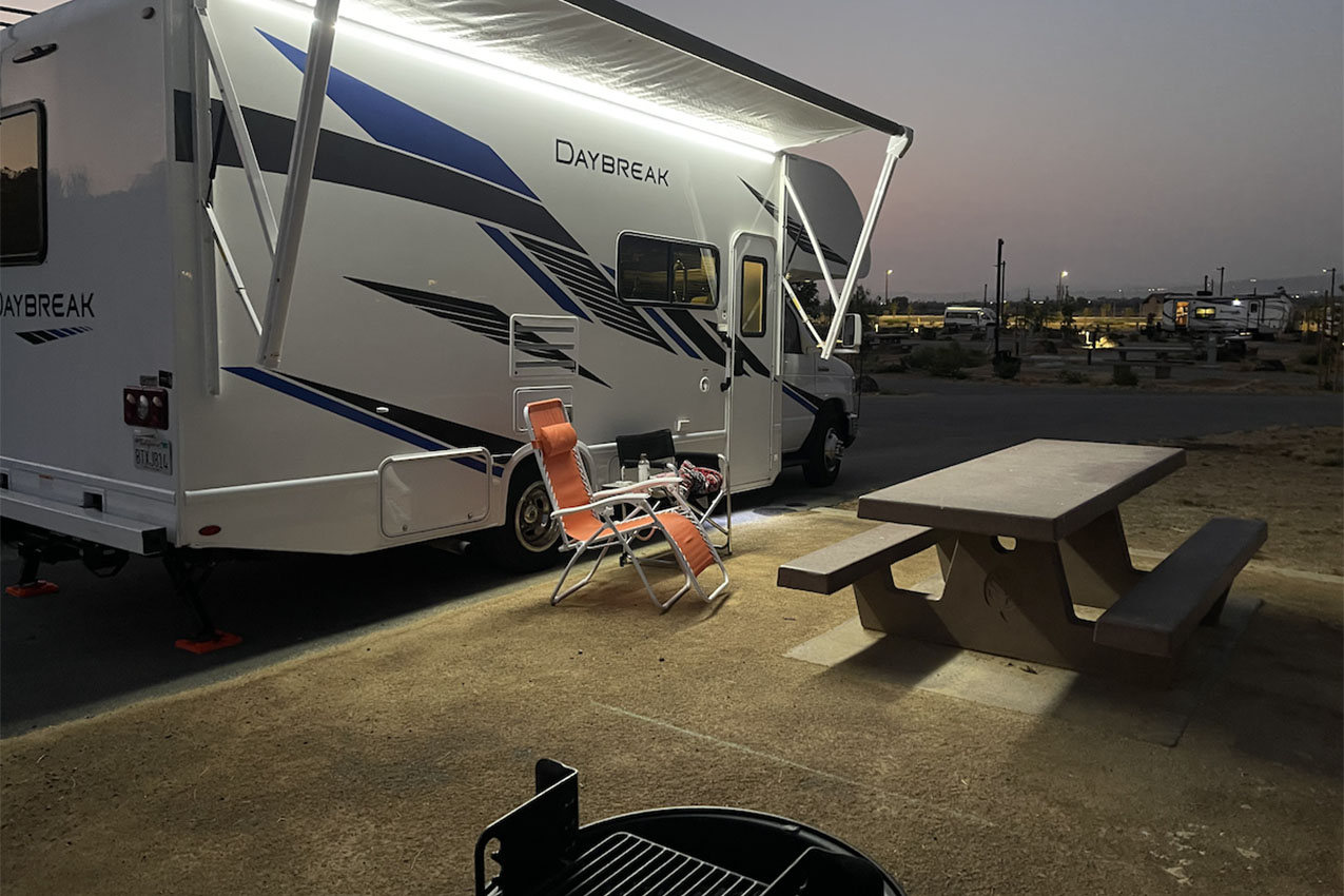 RV with awning and porch light on parked next to a picnic table and fire pit at a campground.