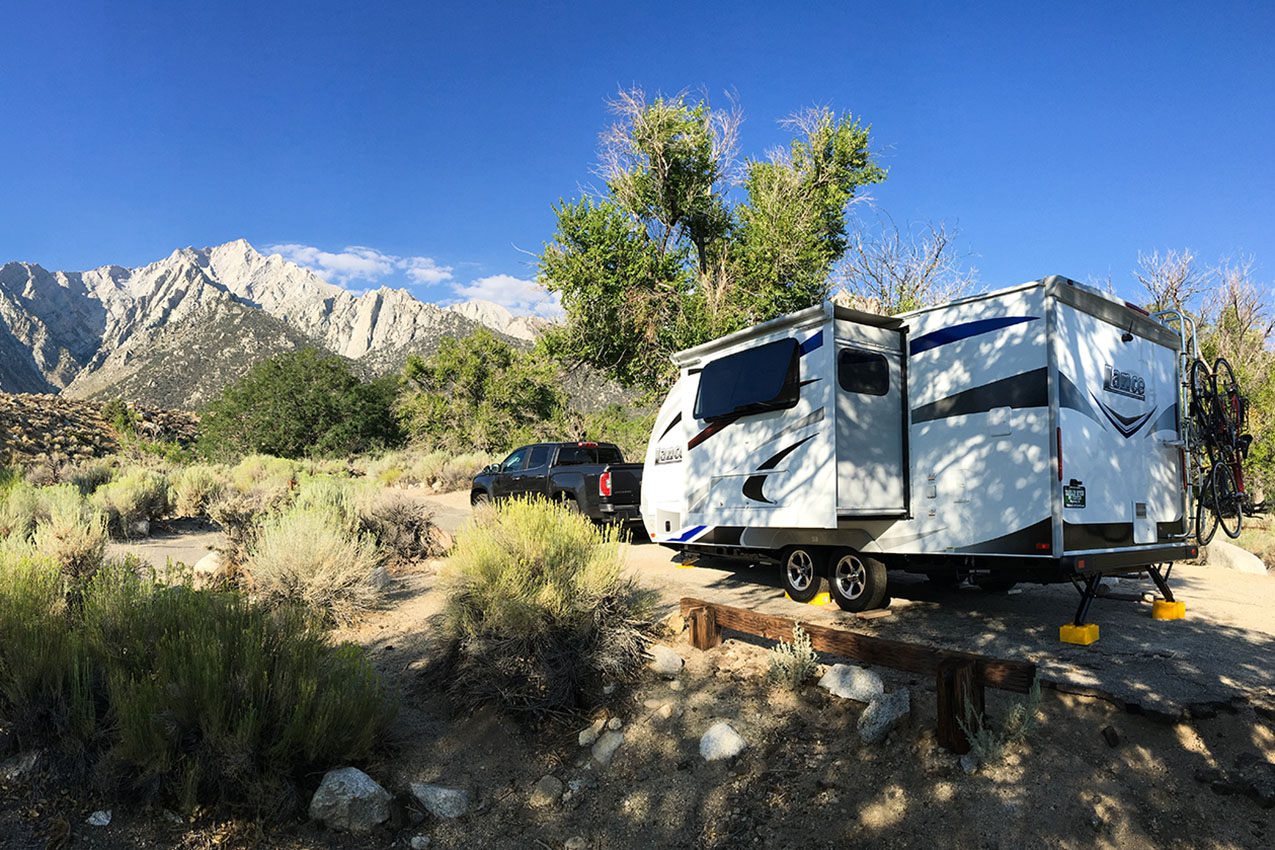 Truck and RV parked in a campground next to a tree in front of large mountains.