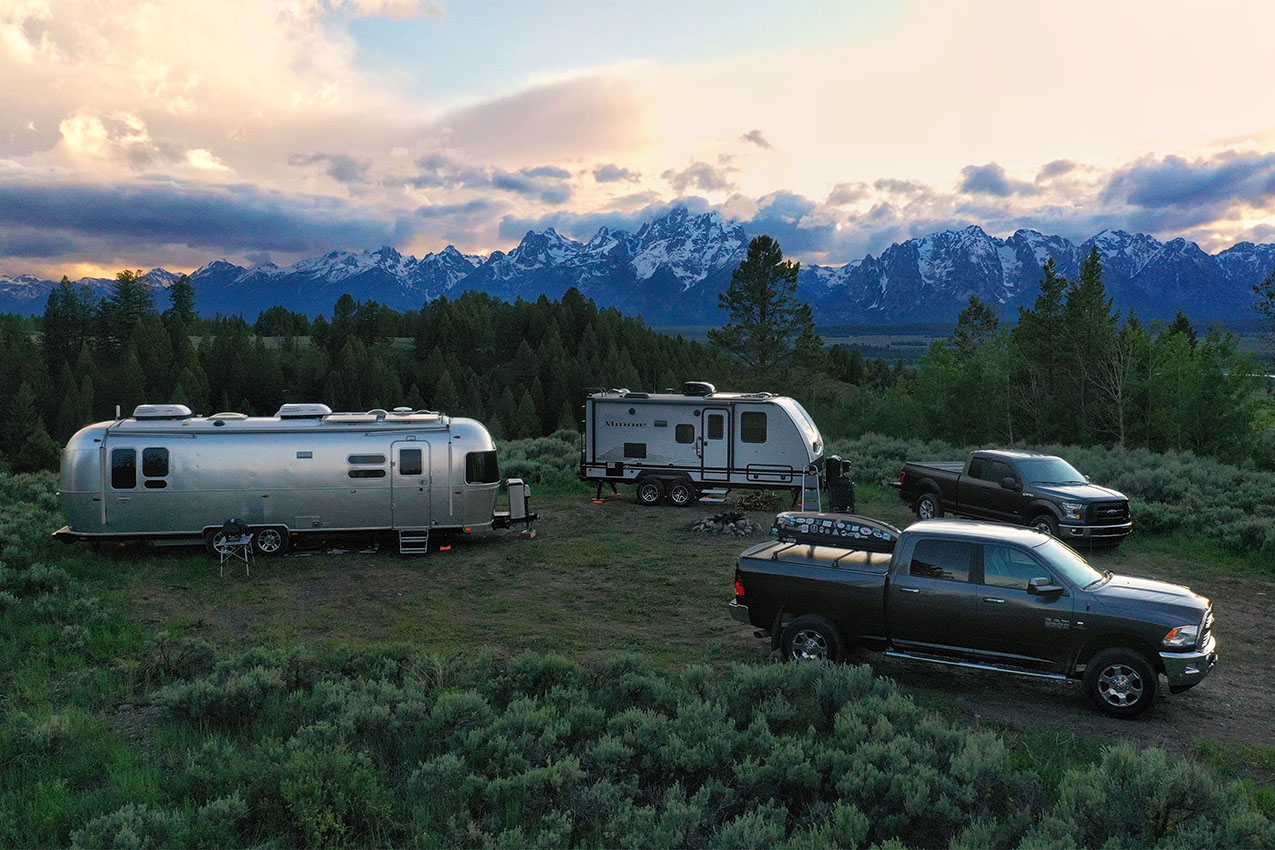 Two RVs and trucks parked in an open grass area overlooking the Teton Mountains at sunset.