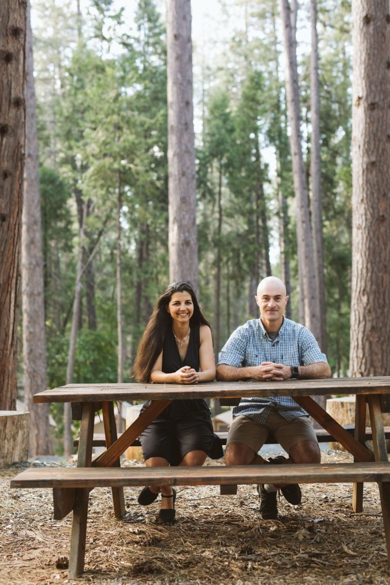 two people sit at a wooden picnic table outside under trees