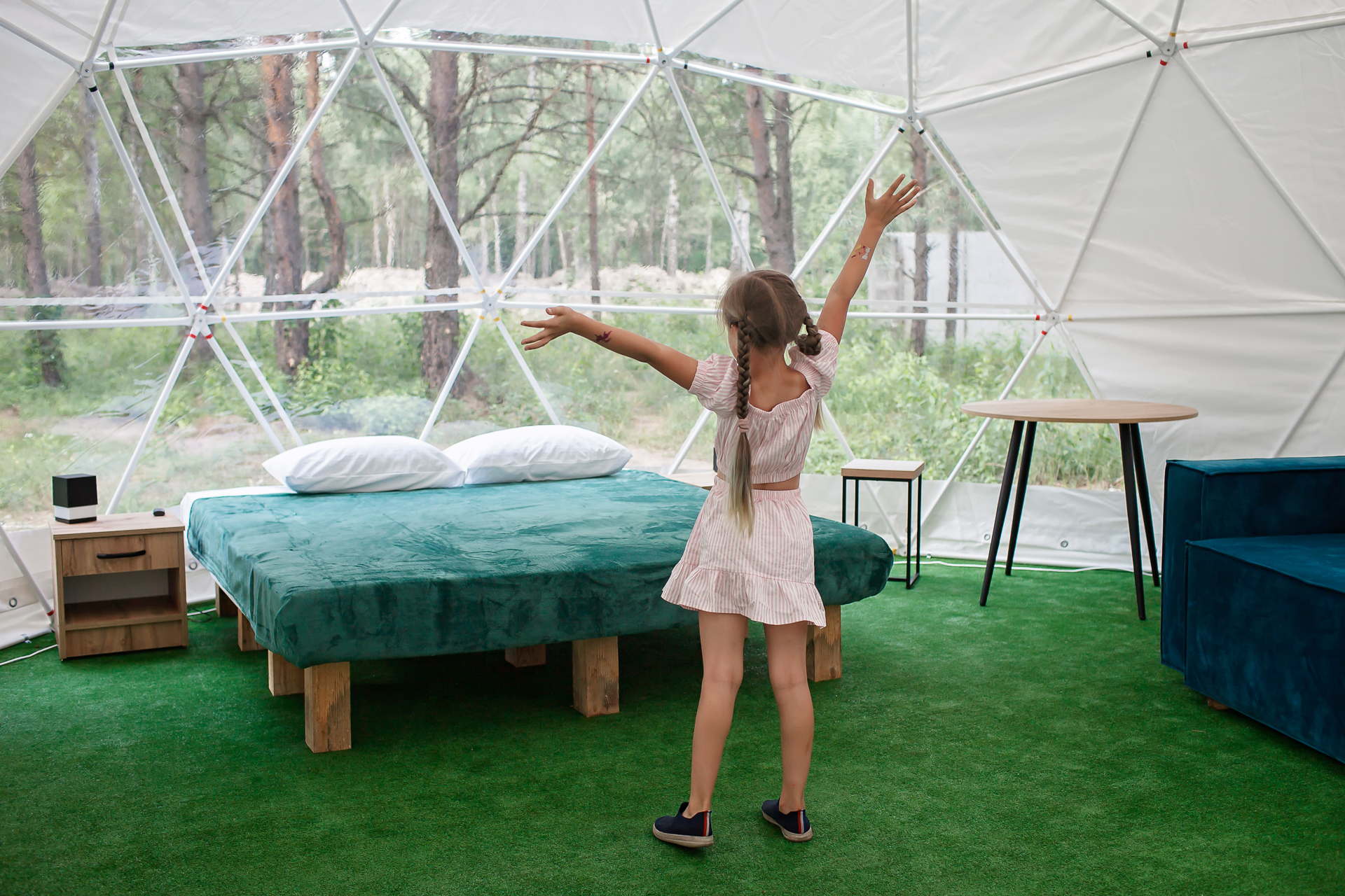 A young girl with her arms outstretched and her back to the camera stands in front of a bed inside a glamping dome