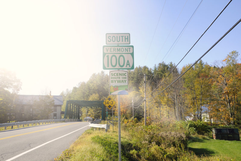 Road sign for Vermont 100A with a bridge and bright sunlight in the background