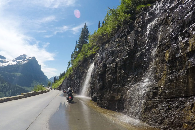 Waterfall flowing onto roadside with motorcyclists and cars on road