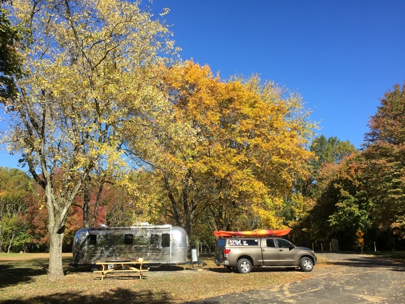An Airstream parked  at a campsite by a truck with a kayak on top.