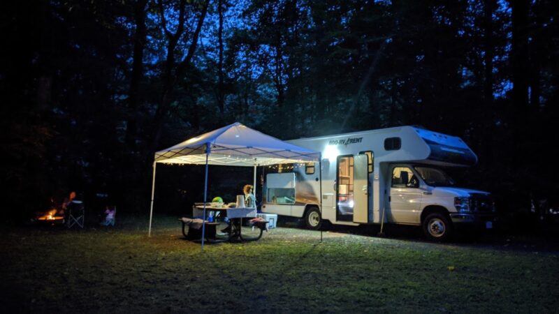 RV at campsite with tent over picnic table with campers enjoying a meal at the campfire
