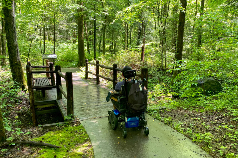 a wheelchair user rolls over a wooden boardwalk on a nature trail through the woods