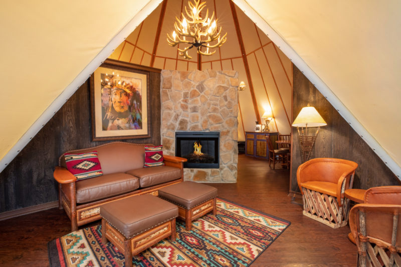 the inside of a teepee with furniture, rugs, an antler light and a stone fireplace