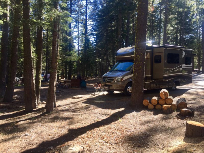 an rv is parked at a wooded campground with a camp chair and a pile of cut logs stacked nearby