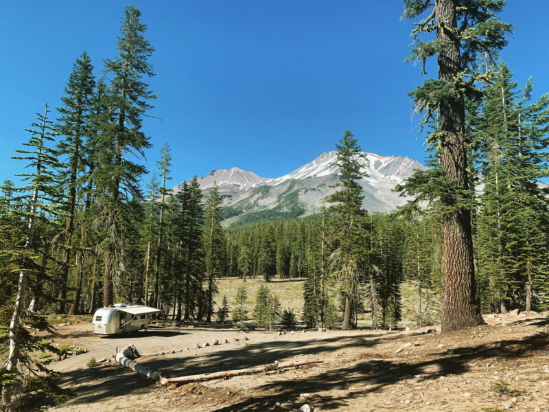 an airstream with an awning is parked at a campground surrounded by pine trees and snowy mountains in the background against a blue sky