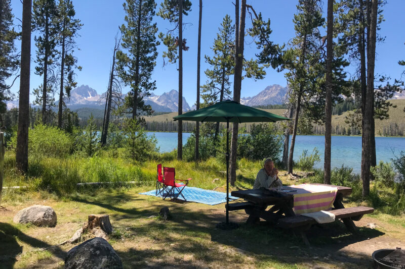 a person sits at a picnic table at a campsite with two camp chairs and an umbrella next to the water with mountains in the background