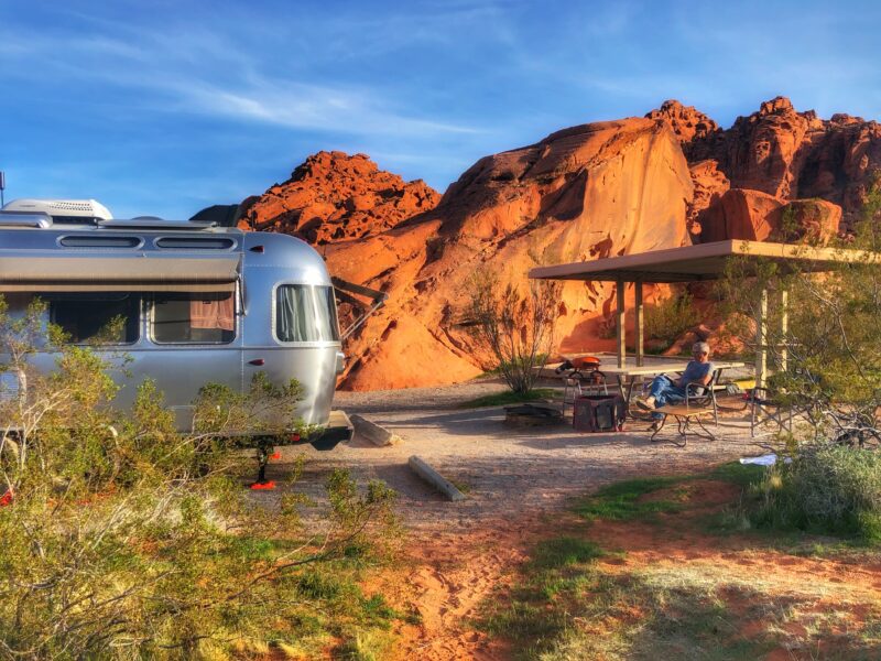 a person sits in a chair next to a silver airstream trailer is parked at a campsite in front of red rocks