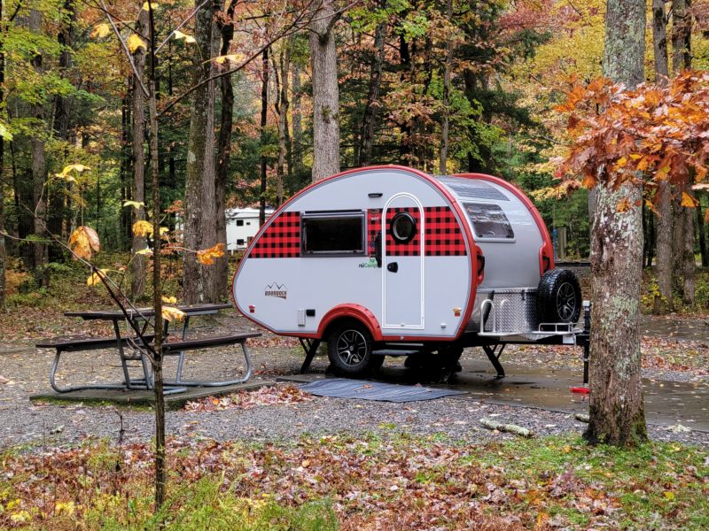 a red striped tear drop trailer is parked at a wooded campsite