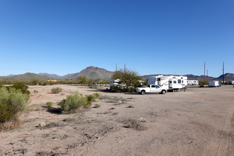 A dirt road through a desert campground with a few RVs and trucks parked
