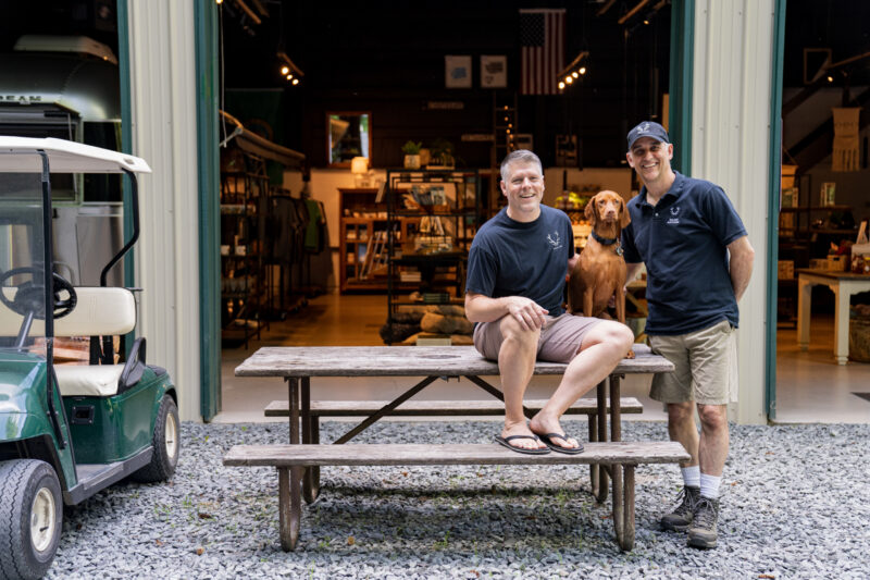 two men and a dog sit at a picnic table in front of a camp store
