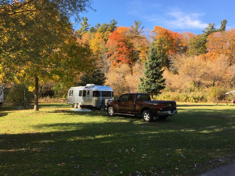 a truck and an airstream trailer parked at a grassy campsite in front of fall foliage