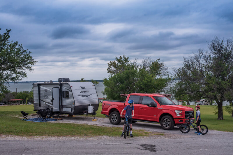 two boys ride bikes at a campsite with an rv and red pickup truck parked nearby