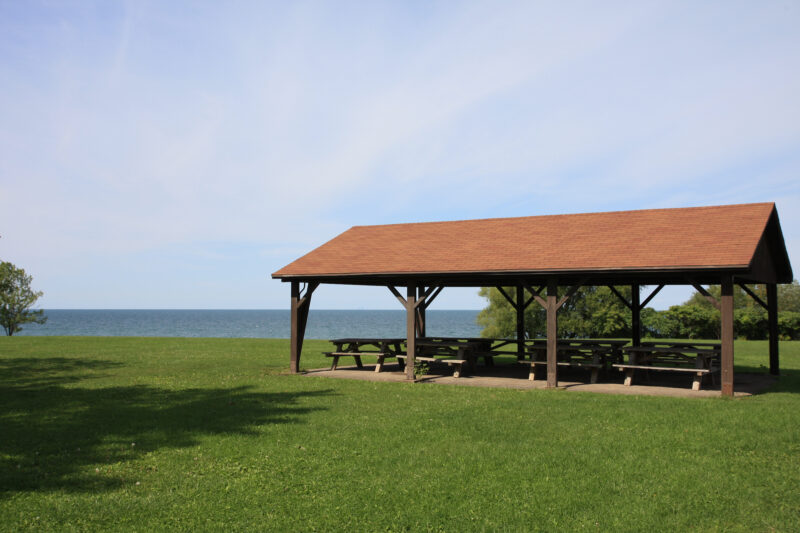 a pavilion with picnic benches sits on a green lawn in front of the water and under a clear blue sky