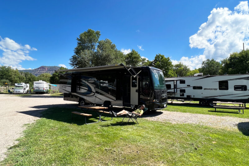RVs are parked at a campsite on grass and gravel under a blue sky with scattered clouds