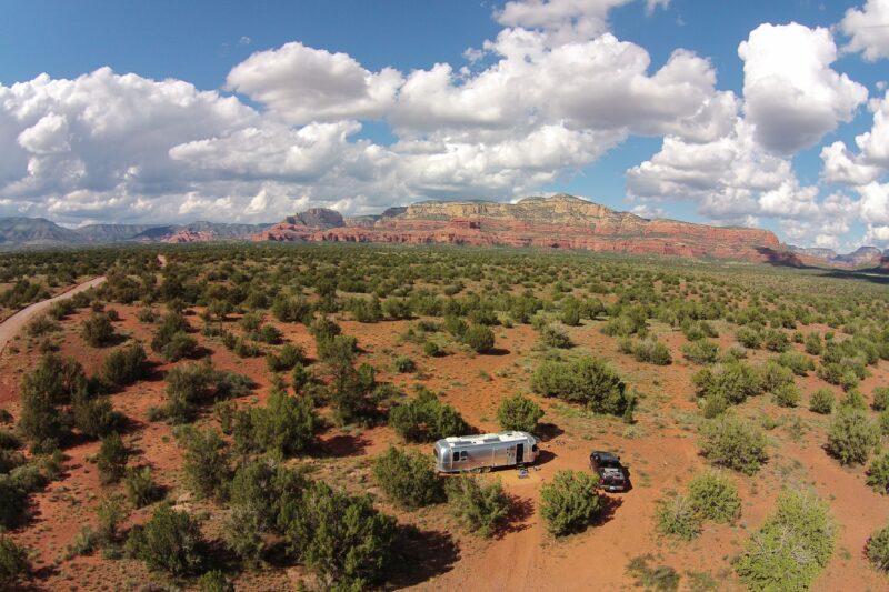 an aerial shot of a desert landscape with a trailer and car parked among scrub brush with red rock formations in the distance